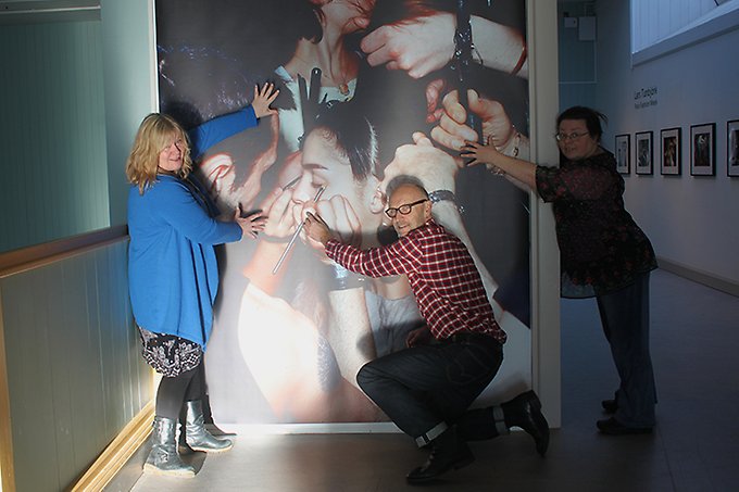 En av de stort uppdragna bilderna på utställningen där en modell stylas av många hjälpsamma händer. Framför bilden showar de tre personer som intervjuas i artikeln