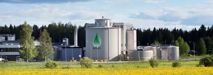 Bild på grå fabrik i sommarlandskap, med logga som visar att fabriken tillverkar biogasbränsle.