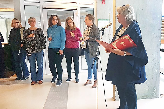 En kvinna står vid en mikrofon och håller tal. Bredvid står en rad kvinnor som åhörare. De håller varsitt glas i händerna. 