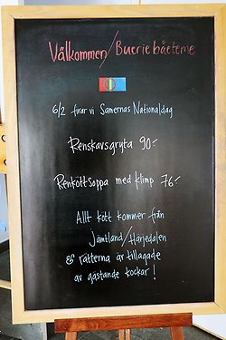 en svart tavla med handskriven meny på. på tavlan står: 6/2 firar vi samernas nationaldag, renskavsgryta 90 kronor, renköttsoppa med klimp 76 kronor, allt kött kommer från jämtland/härjedalen och rätterna är tillagade av gästande kockar