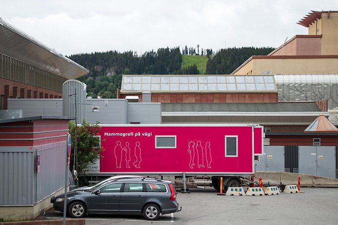 En stor vagn med rosa väggar och texten Mammografi på väg står uppställd på en parkering utanför Östersunds sjukhus. Framför vagnen står några personbilar parkerade. 