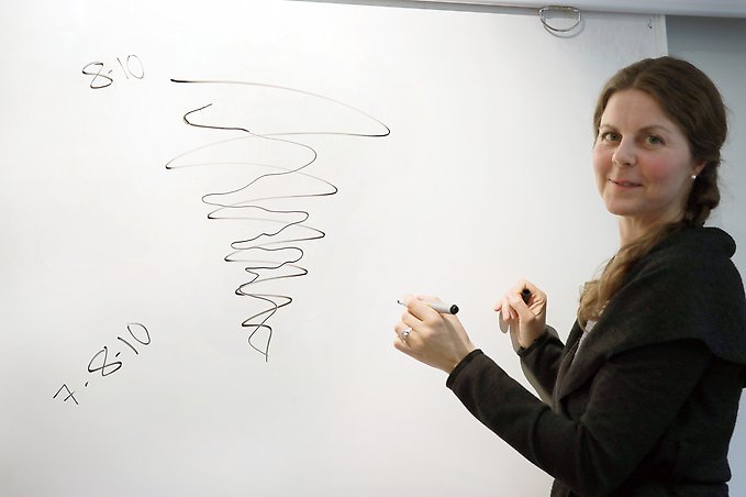 en kvinna står framför en whiteboard med en penna i handen och tittar in i kameran