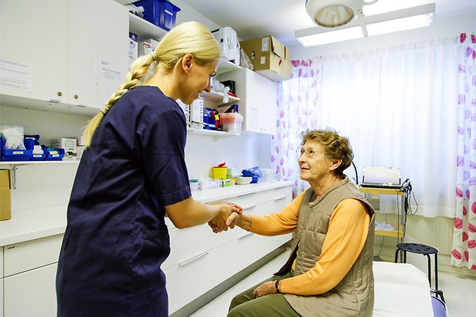 en blond kvinna med hårfläta och mörkblå vårdkläder står framför, och håller handen på en äldre kvinna som sitter på en bädd i ett undersökningsrum