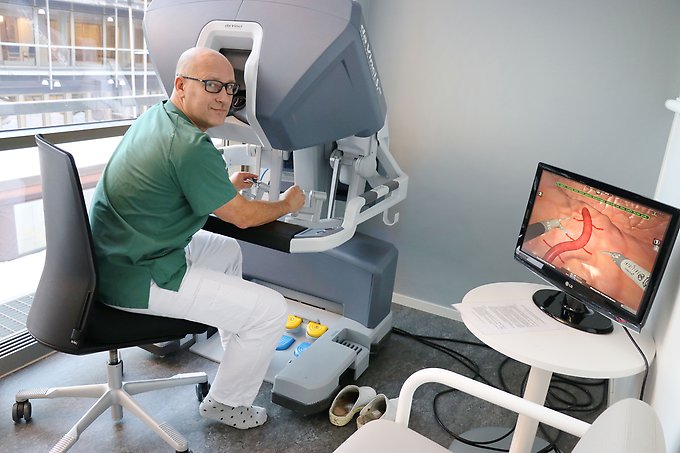 en manlig läkare sitter vid en robotkirurgimodul och kör ett övningsprogram som syns på en datorskärm på ett bord bredvid modulen