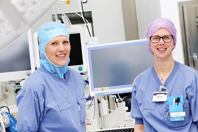 en kvinnlig sjuksköterska och en kvinnlig undersköterska, båda iklädda operationskläder, står bredvid varandra framför en datorskärm i en operationssal