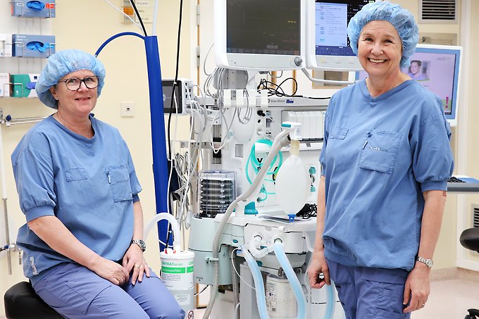 två personer klädda i blå vårdkläder och hårnät intill en anestesiapparat