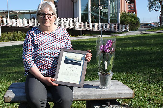 En kvinna sitter på en träbänk i solen. I händerna håller hon i ett diplom, och bredvid henne på bänken står en orkidé i kruka