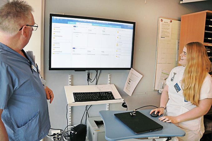 en man och en kvinna står och tittar på en stor datorskärm på väggen, kvinnan arbetar på skärmen med hjälp av en datormus