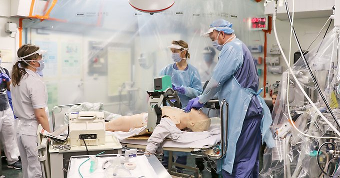 tre personer står runt en sjukhussäng på vilken det ligger en övningsdocka, i bakgrunden syns en person som står bakom ett plastskynke