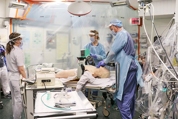 tre personer står runt en sjukhussäng på vilken det ligger en övningsdocka, i bakgrunden syns en person som står bakom ett plastskynke