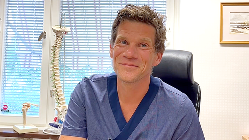 En läkare i blå vårdkläder sitter bakom ett skrivbord.