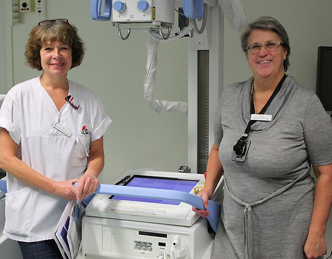 Två kvinnor står på var sin sida om en portabel röntgenmaskin, de ler mot kameran