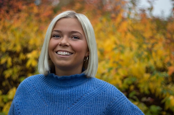 Närbild utomhus på en leende person med blå tröja och axellångt, ljust, rakt hår som står framför en buske med gula löv.