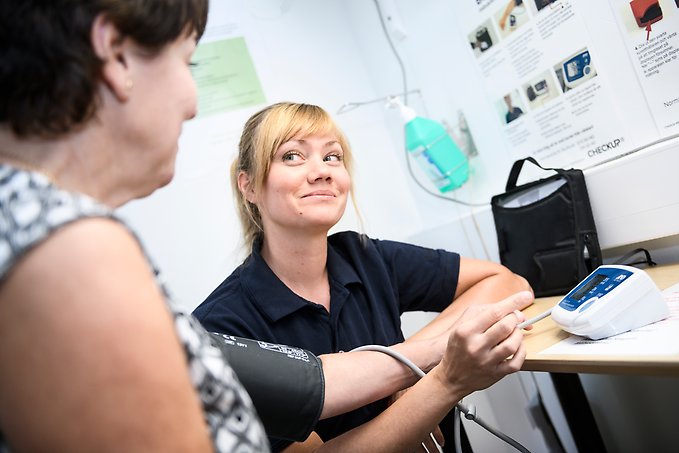 en sjuksköterska sitter på huk bredvid en patient som sitter vid ett skrivbord med en blodtrycksmanschett på överarmen.