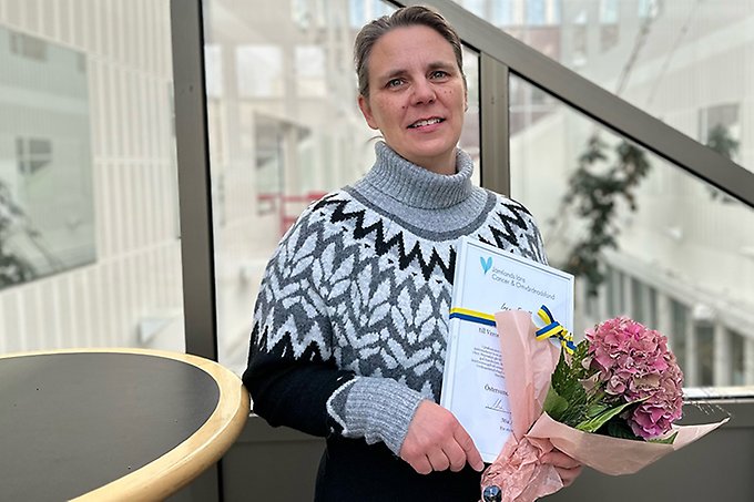 Person står vid ett runt bord med blommor och ett stipendium i händerna. Ler mot kameran.