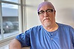 En glasögonprydd person klädd i blå operationskläder och hätta på huvudet står vid ett fönster och tittar in i kameran.