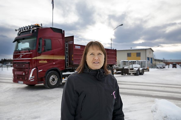 En person med axellångt mörkt hår och svart jacka står på en trottoar och tittar in i kameran. I bakgrunden syns en röd lastbil med släp som ska korsa vägen bakom personen. 