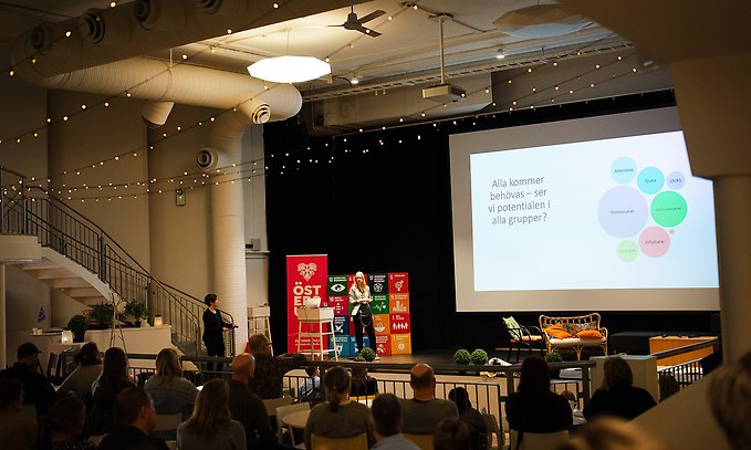 Talare på scen vid en presentationsskärm, med en publik av åhörare nedanför. På presentationsskärmen syns texten: "Alla kommer behövas – ser vi potentialen i alla grupper?"