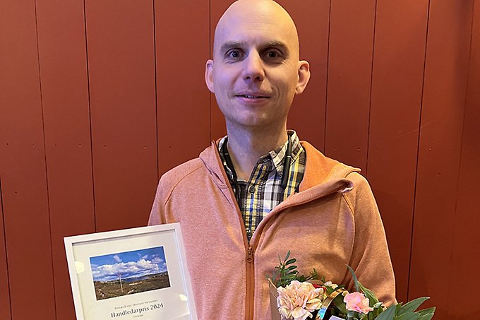 En person står mot en röd vägg med blombukett och diplom i händerna. Ser glad ut.