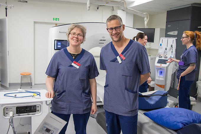 Två personer står framför en datortomografiapparat, i bakgrunden står två personer och pratar med varandra.