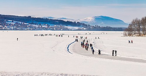 Landskapsbild på Oviksfjällen i bakgrunden, Annersias glesbygd i förgrunden och isen över Storsjön där massa människor åker skridskor på uppskottade banan Medvinden. Bilden ska symbolisera Jämtland Härjedalens invånare, urbana delar, landsbygd och natur.