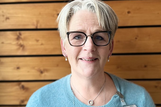Bild på leende person med kort blont hår och glasögon. Iklädd ljudblå tröja. Bakgrund trävägg.