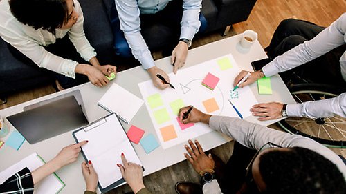 Flera personer sitter runt ett mötesbord och samarbetar. Man ser deras händer och skjortklädda ärmar måla en gemensam skiss och märka upp den med post-it-lappar.