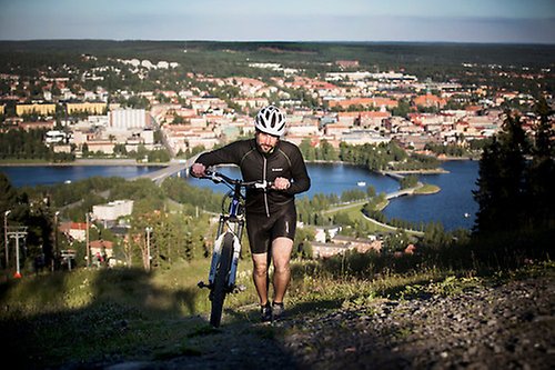 En man går upp för en backe med en cykel. Bakom honom syns en stad.