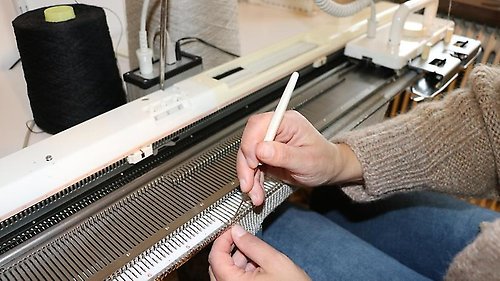 Närbild på ett par händer som jobbar med nål och garn vid en handstickningsmaskin.